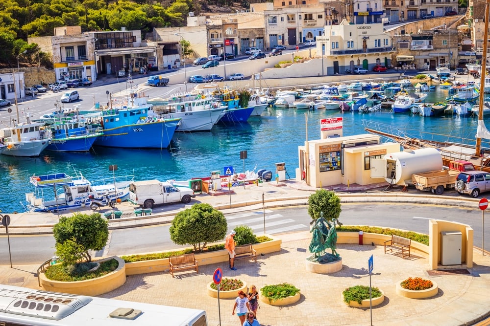  Beautiful View of Port of Mgarr on the Small Island of Gozo, Malta. A vibrant harbor with things to do in Malta, boats docked along the pier, surrounded by buildings and a statue, with people walking about on a sunny day.