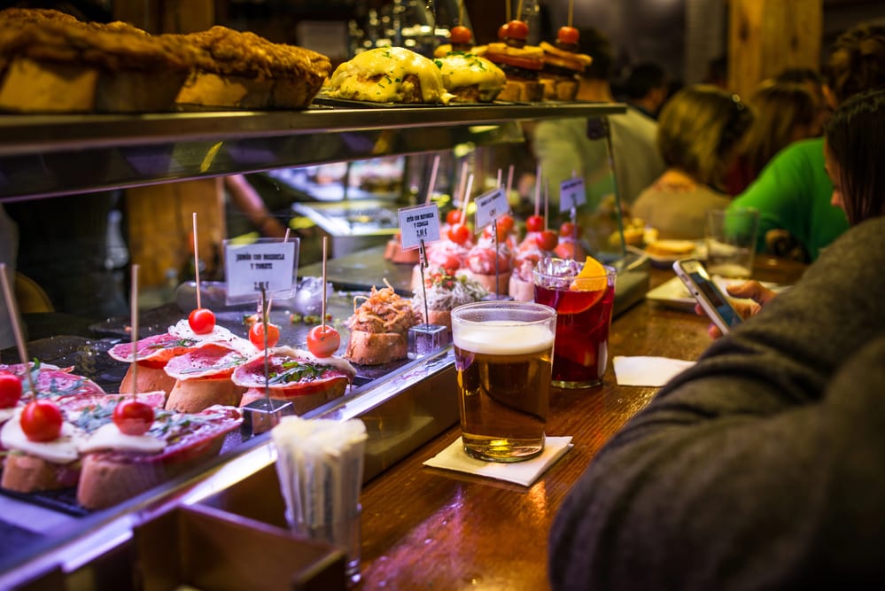 A vibrant tapas bar in Madrid with an array of snacks, a glass of beer, and patrons in the background.