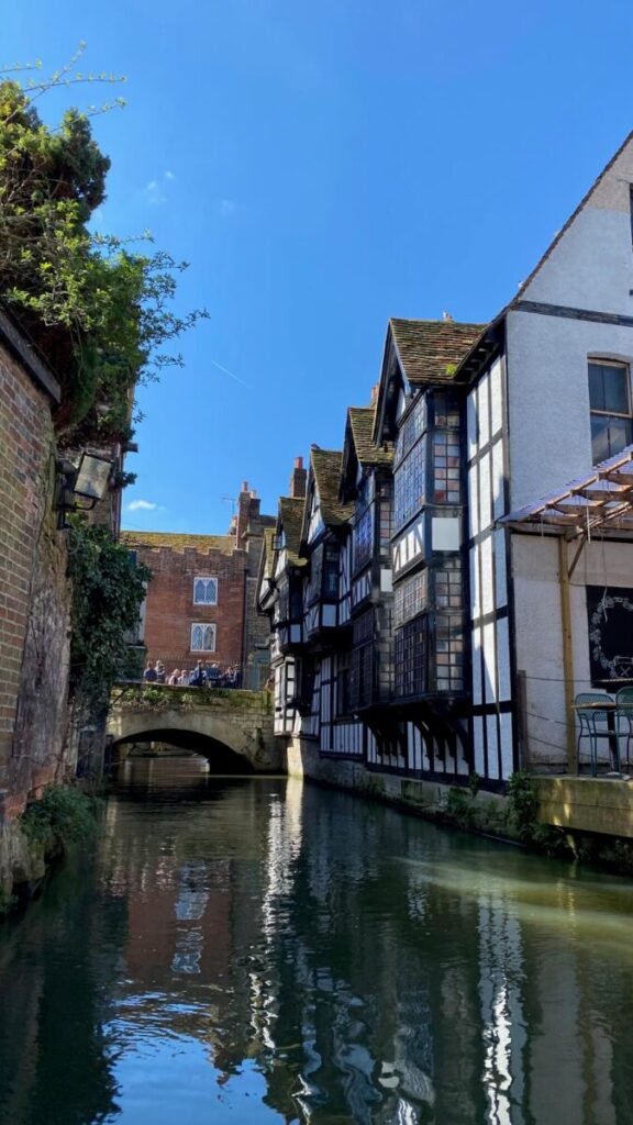 Half-timbered buildings overlooking a narrow canal with Canterbury boat trips under a clear blue sky.