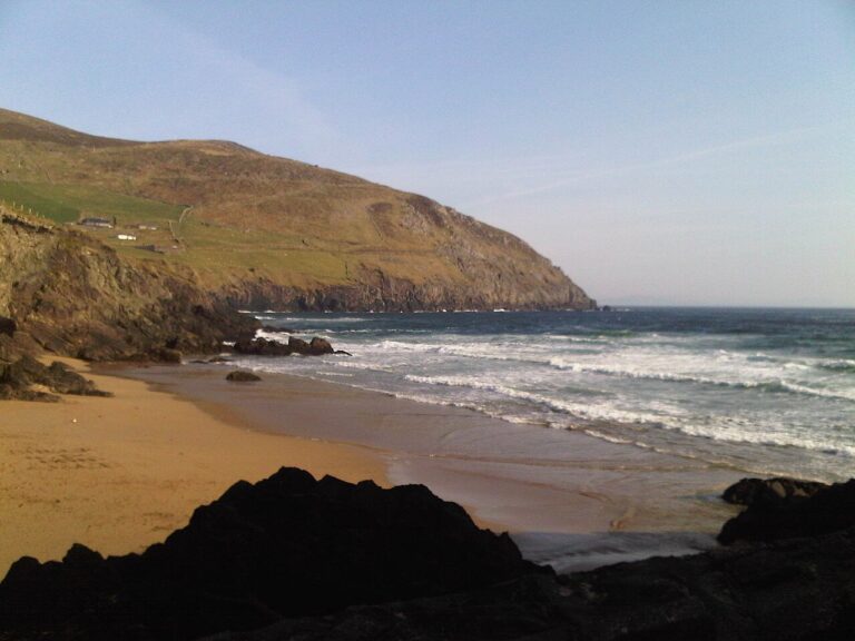Waves crash on a sandy beach below the green hills of Irelands Dingle Peninsula.