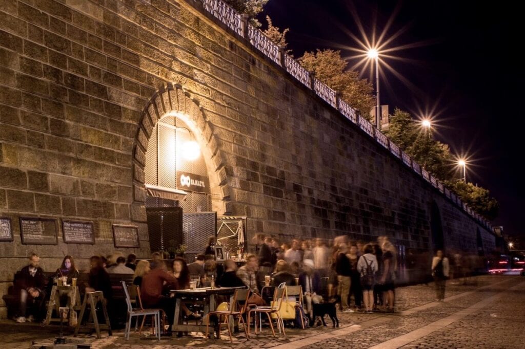 Nighttime scene at a bustling outdoor cafe in Prague, located under a large stone bridge, with patrons seated at tables and others standing, illuminated by warm lights.