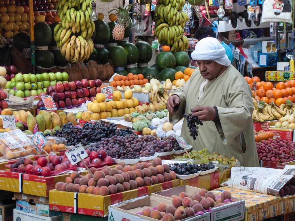 A man in a white head covering and tan robe examines a bunch of grapes at a fruit stand filled with various produce.
