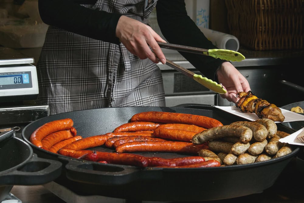 Person cooking various sausages and skewered meat on a large frying pan, using green tongs, inspired by a recent visit to Prague.