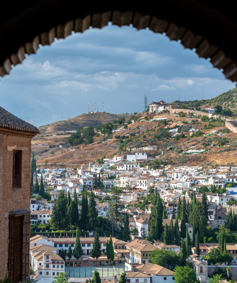 A view of a hillside town with white buildings and cypress trees framed by a stone archway.