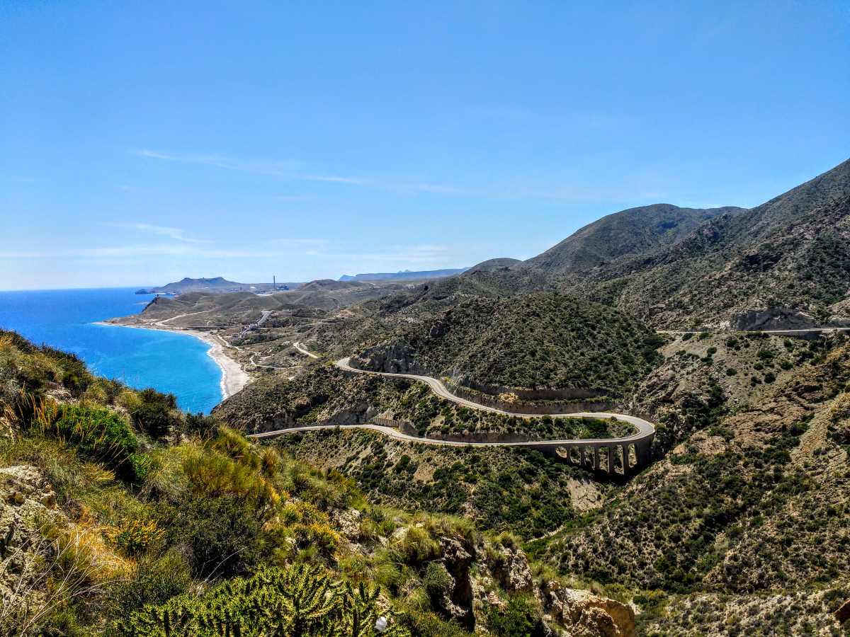 A winding road curves through arid, hilly terrain overlooking a blue coastline under a clear sky.