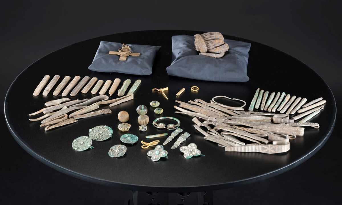 A collection of ancient metal artifacts, including strips, brooches, and beads, is arranged on a black circular table.