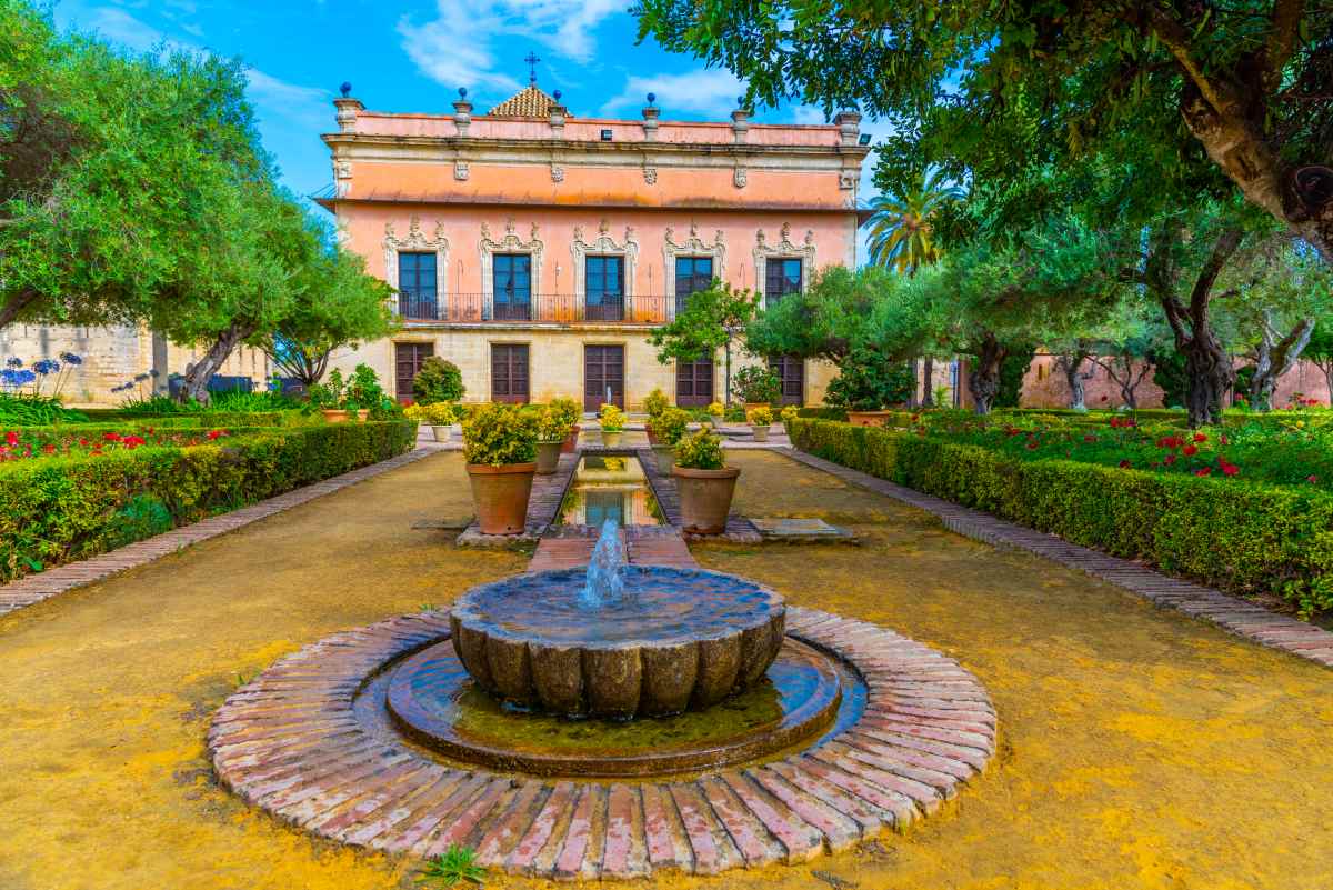 A stone fountain sits in a garden path leading to a multi-story building with a peach-colored facade and dark windows.