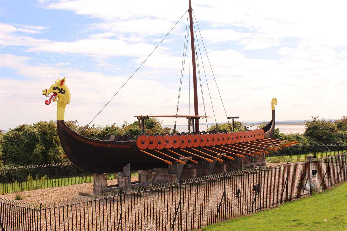 A wooden ship model with a dragon-headed prow and oars sits on a stone base behind a metal fence.
