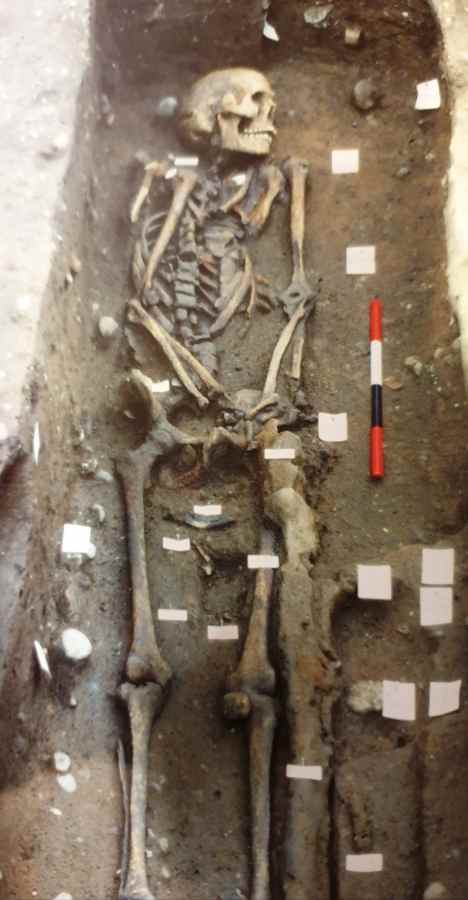 A human skeleton lies in a narrow dirt trench alongside a red, white, and black measuring scale and several small markers.