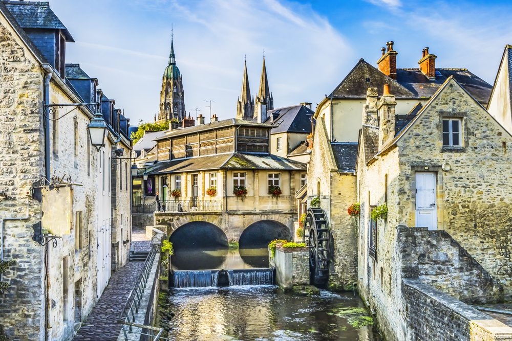 A stone building with a water wheel sits beside a canal in a town with historic architecture and church spires.