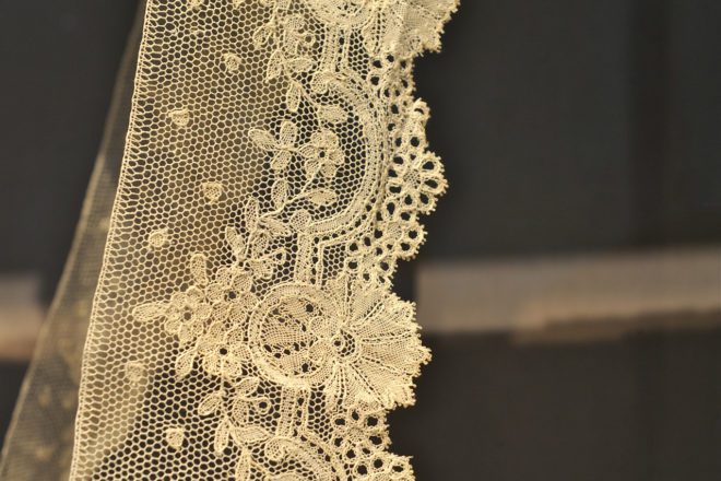 A strip of cream-colored lace features a hexagonal mesh pattern with intricate floral and scalloped edge embroidery.