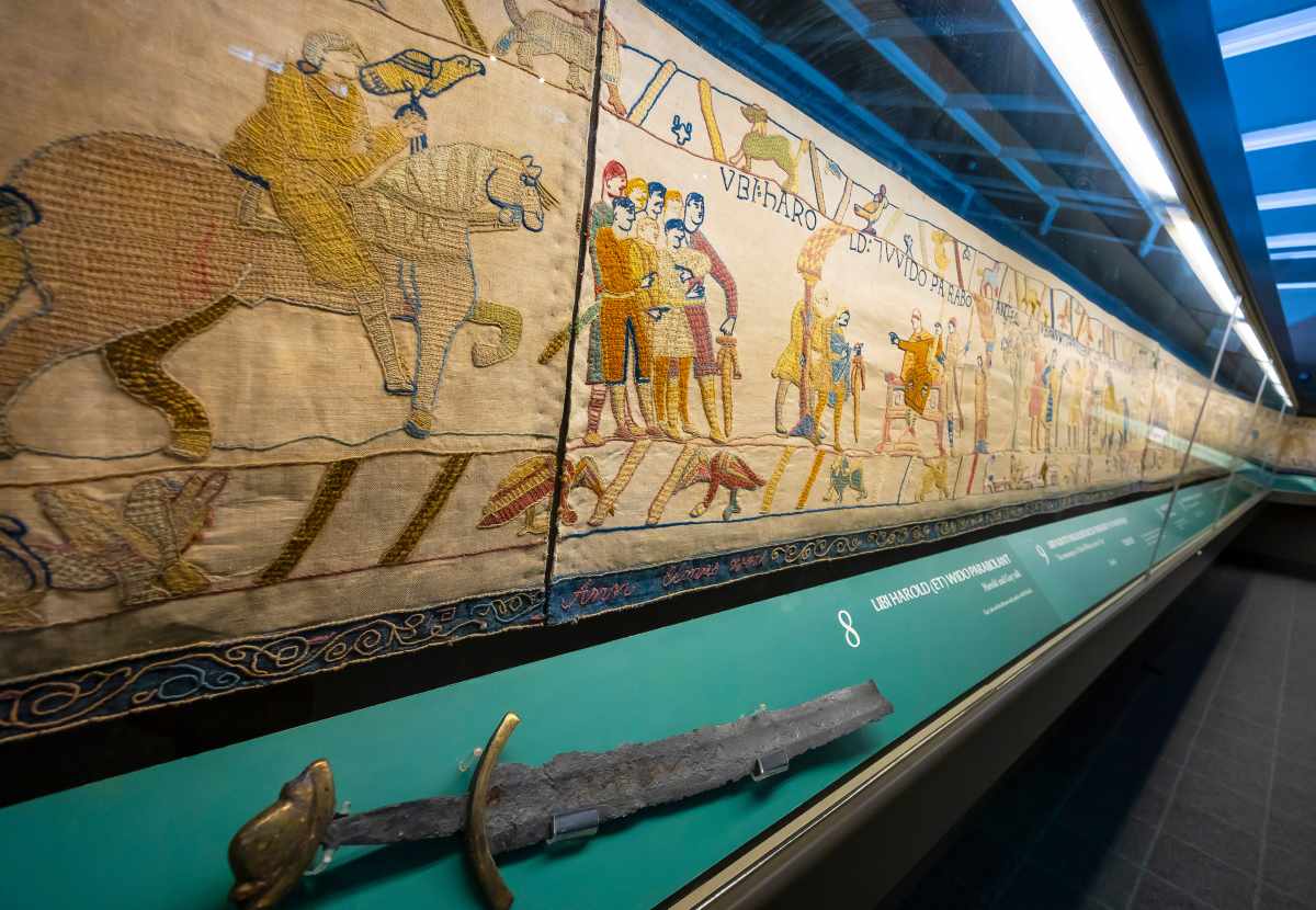 An embroidered tapestry depicting historical figures and animals is displayed behind glass above an antique sword.