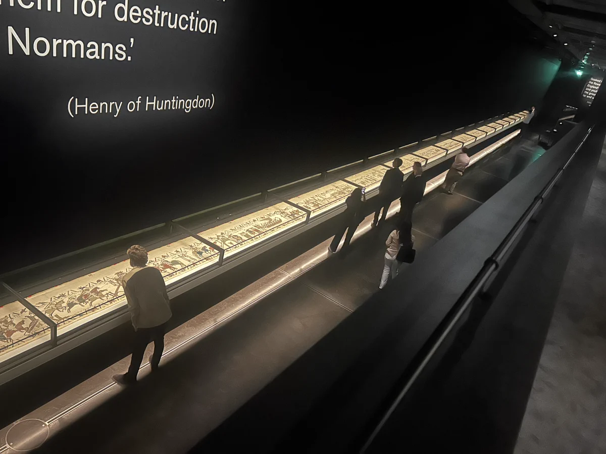 Visitors walk along a long, illuminated display case containing a detailed historical tapestry in a dark museum gallery.