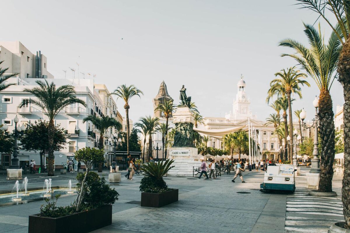 People walk through a sunny plaza featuring a central statue, palm trees, and surrounding buildings.