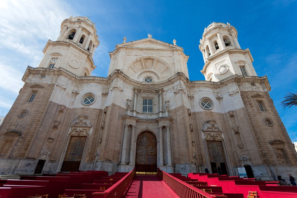 A large stone cathedral with two towers stands behind a red-carpeted ramp and seating area under a clear blue sky.