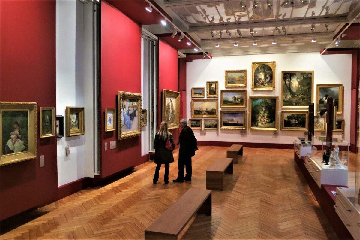 Two people stand in an art gallery with many framed paintings displayed on red and white walls above a wooden floor.