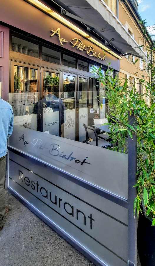 The exterior of a restaurant named Au Ptit Bistrot features a glass-enclosed patio with outdoor seating and greenery.