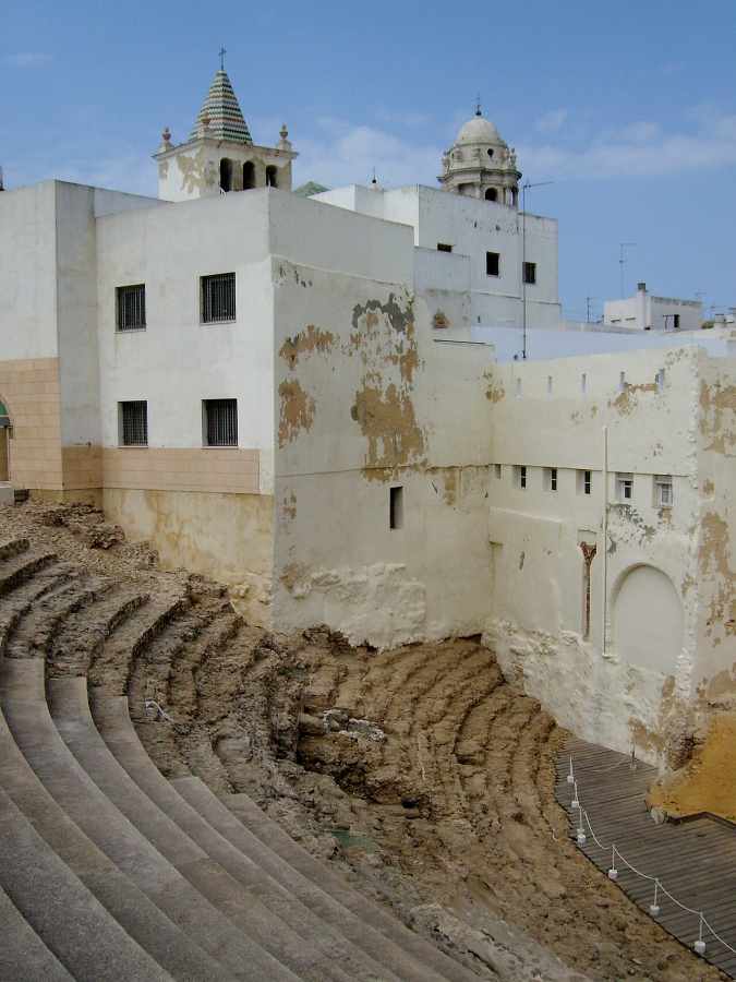 Ancient stone amphitheater ruins sit adjacent to white buildings with a tiled steeple and a dome under a blue sky.