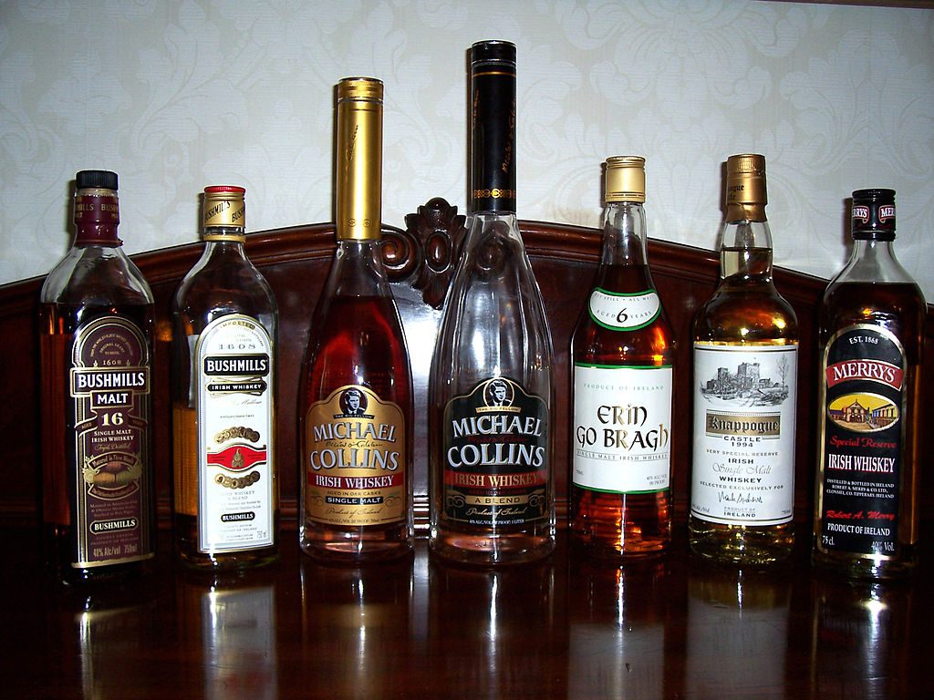 Seven bottles of Irish whiskey are arranged in a row on a dark wooden surface against a patterned wallpaper background.