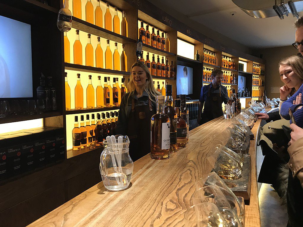 A woman and a man in aprons stand behind a wooden bar lined with many bottles of amber liquid and tasting glasses.