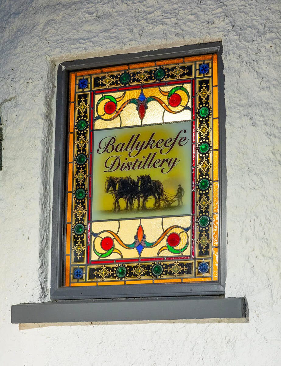 A stained glass window features the text Ballykeefe Distillery above an illustration of a person plowing with two horses.
