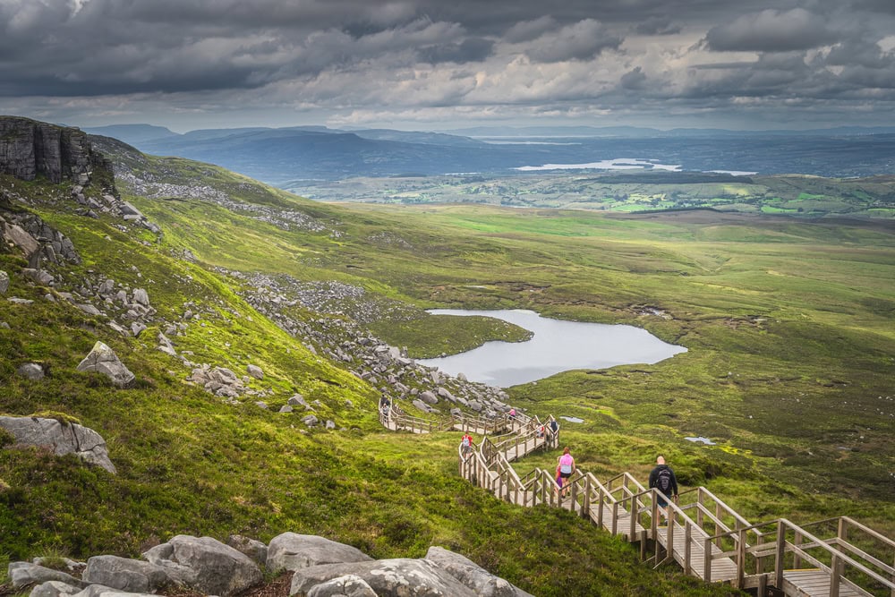 People enjoying a walk on steep stairs of wooden boardwalk on the bank of the Cuilcagh Mountain with a view on lake and valley below, Northern Ireland