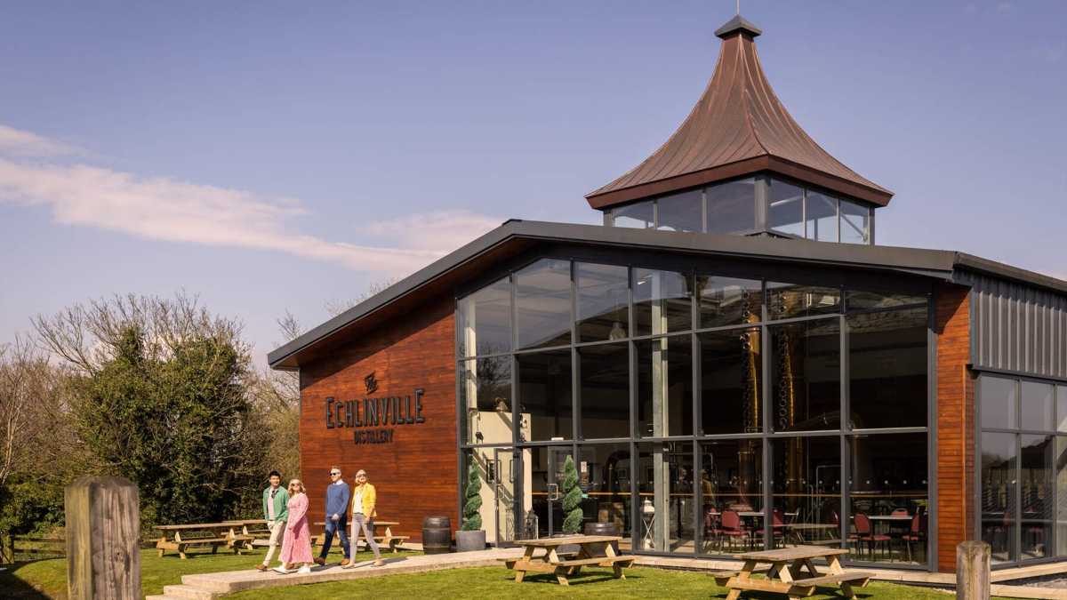 Four people walk on a path in front of a modern wooden building with a glass facade and a pointed roof.