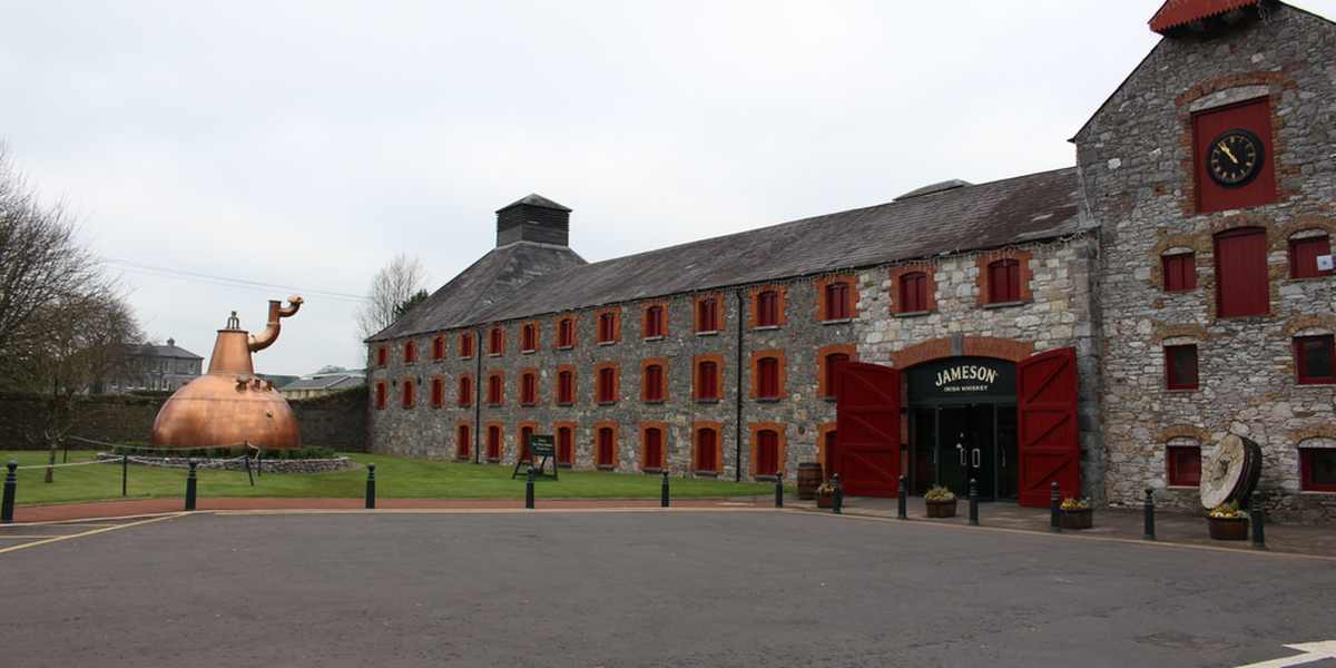 A stone building with red-trimmed windows features a large copper still on the lawn and an open entrance marked Jameson.