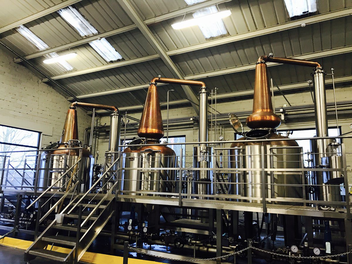 Three large copper and stainless steel industrial stills stand on a metal platform inside a warehouse.