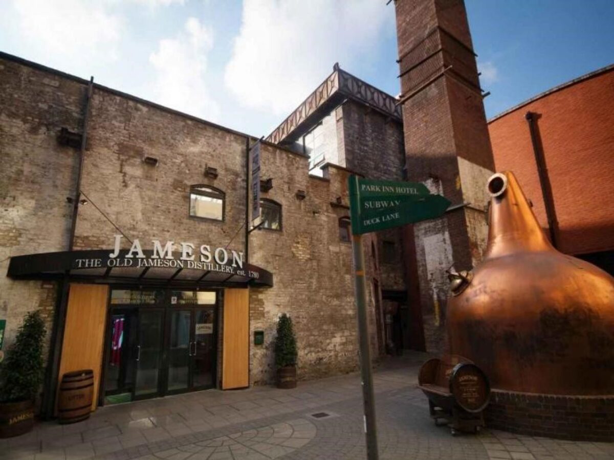A brick building with a Jameson sign stands next to a large copper still and a green directional sign.