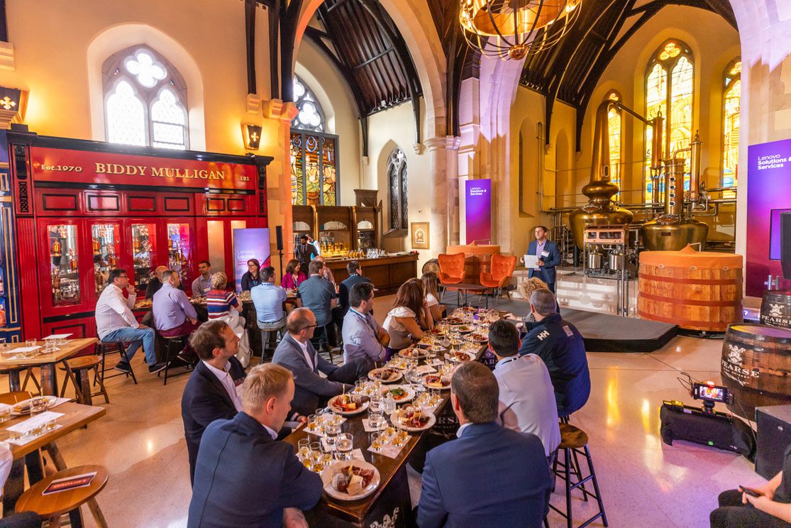 People sit at long tables in a large, vaulted room with stained glass windows and copper distilling equipment.