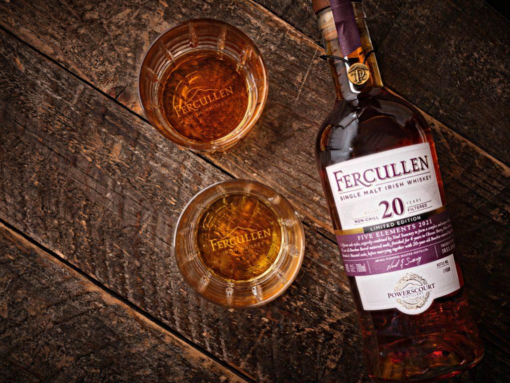 A bottle of Fercullen 20-year-old Irish whiskey sits next to two glasses of whiskey on a dark wooden surface.