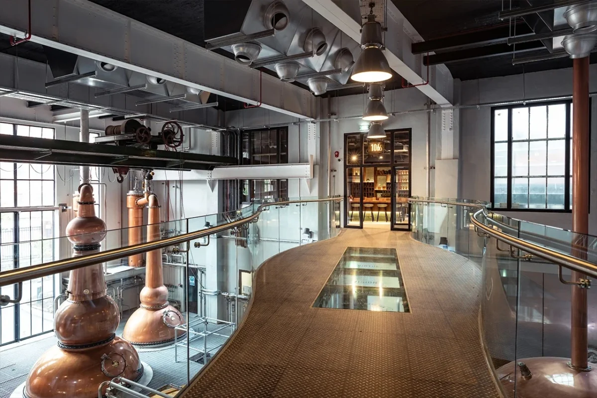 A metal walkway with a glass floor overlooks large copper stills in an industrial-style building with high ceilings.