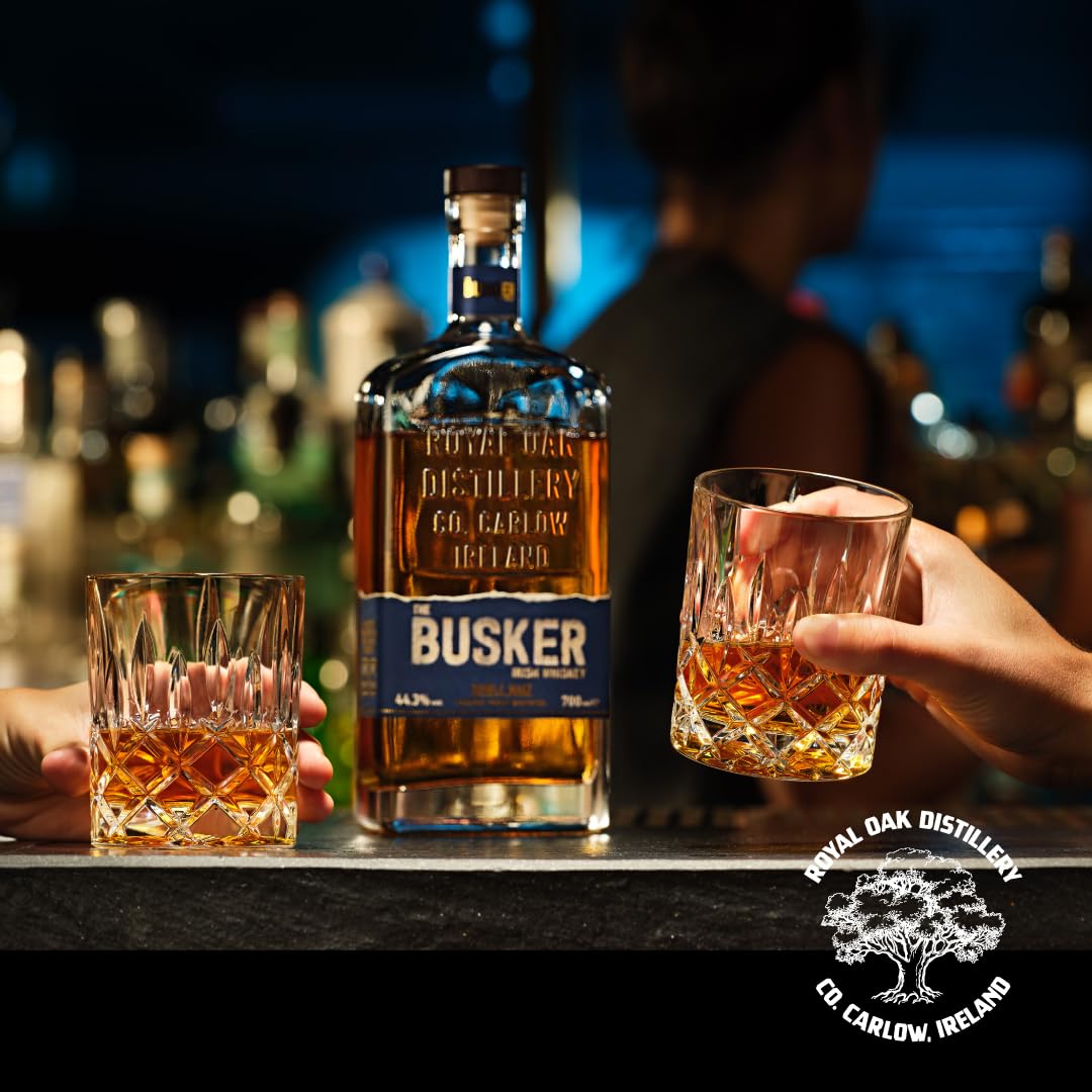 A bottle of The Busker Irish Whiskey stands between two hands holding crystal glasses filled with amber liquid.