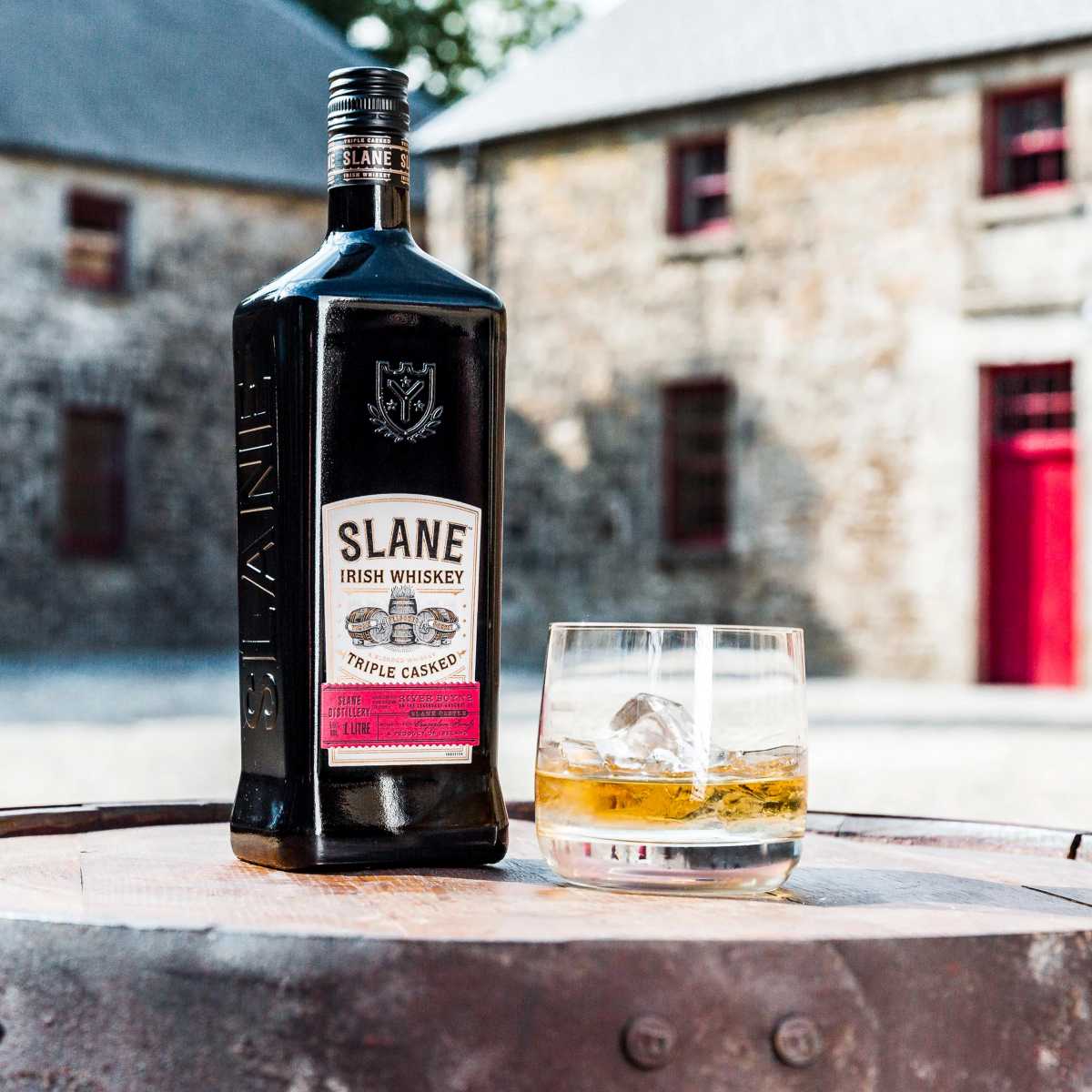 A bottle of Slane Irish Whiskey and a glass with ice sit on a wooden barrel in front of a stone building.