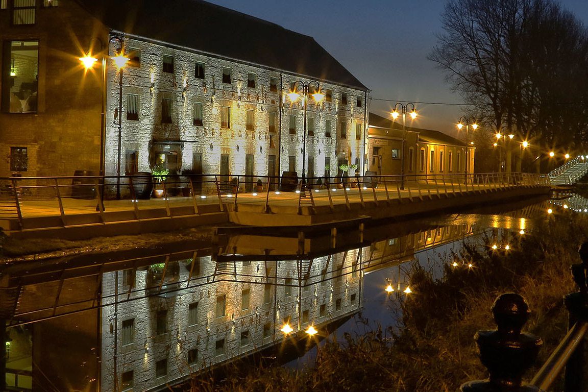 A stone building with many windows is illuminated at night along a canal, reflecting in the calm water.