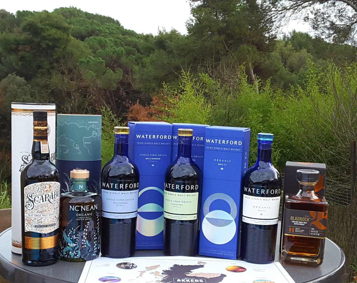 Several bottles of whiskey and their boxes are arranged on a round table outdoors against a background of green trees.