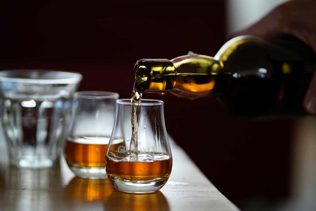 Amber liquid is poured from a bottle into a small glass on a wooden surface next to other empty glasses. Whiskey Distillery tours