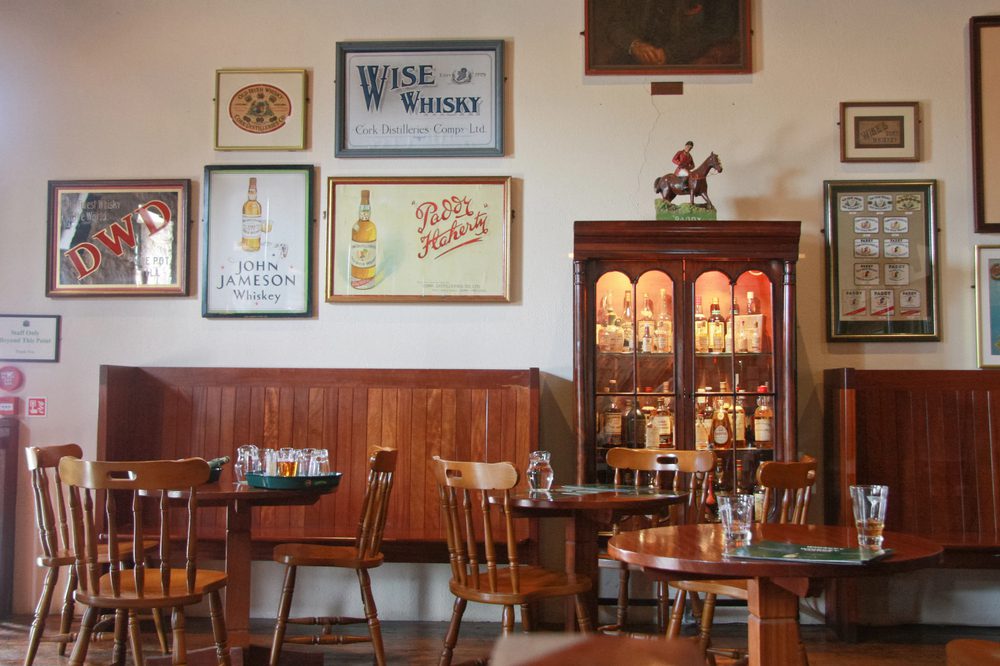 A room features wooden tables and chairs, a glass-fronted liquor cabinet, and various vintage whiskey advertisements on