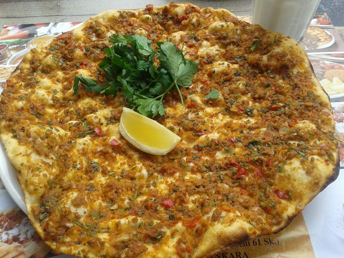 A round, thin flatbread topped with minced meat, herbs, and red peppers, garnished with fresh parsley and a lemon wedge.