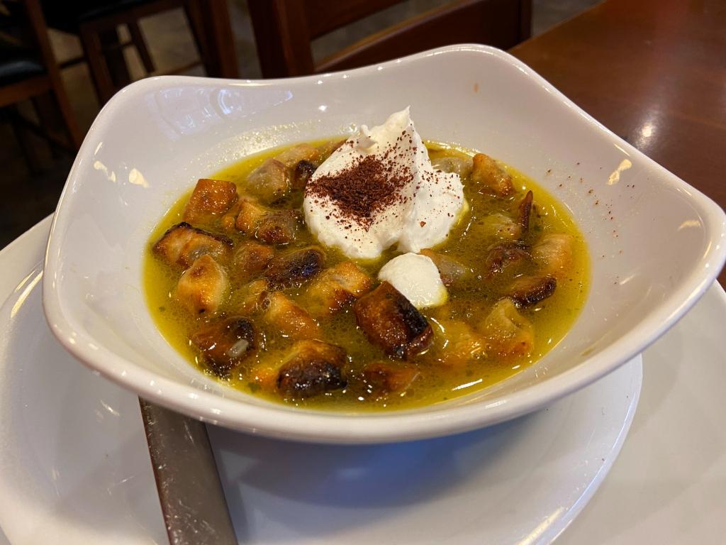A white bowl contains browned, bite-sized pieces in a yellow broth topped with a dollop of cream and a dusting of spice.