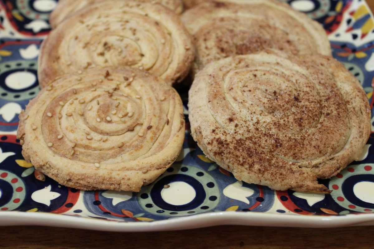 Several round, spiral-shaped cookies topped with sesame seeds and cinnamon rest on a patterned plate.