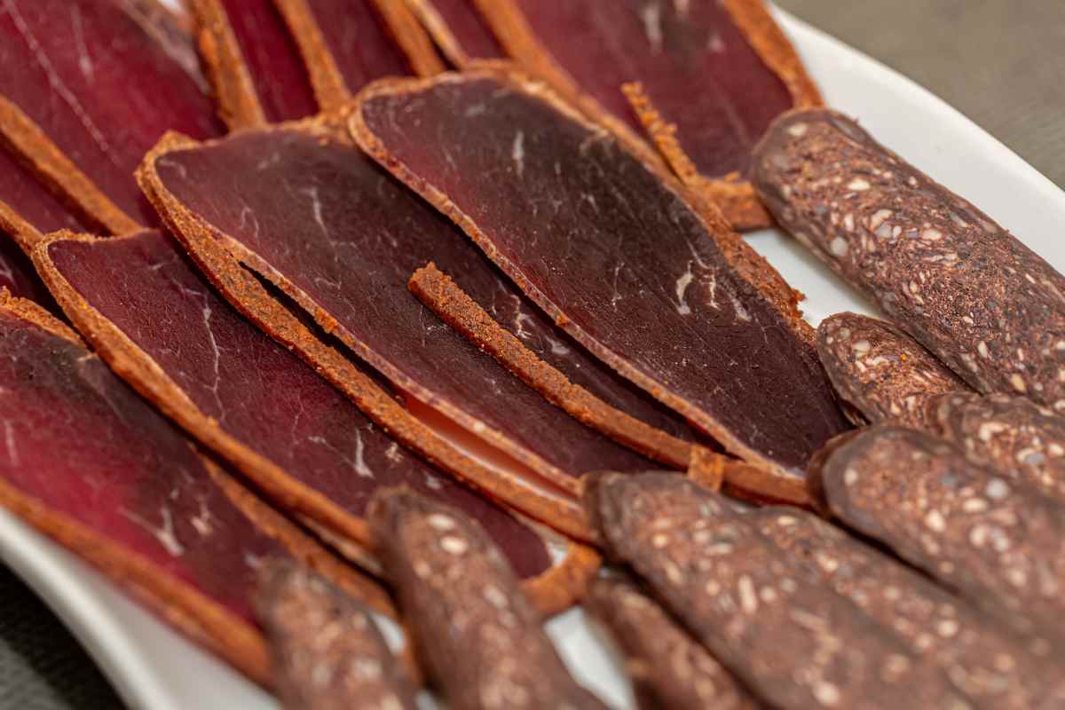 Thin slices of cured meat and pieces of dark, textured sausage are arranged on a white plate.