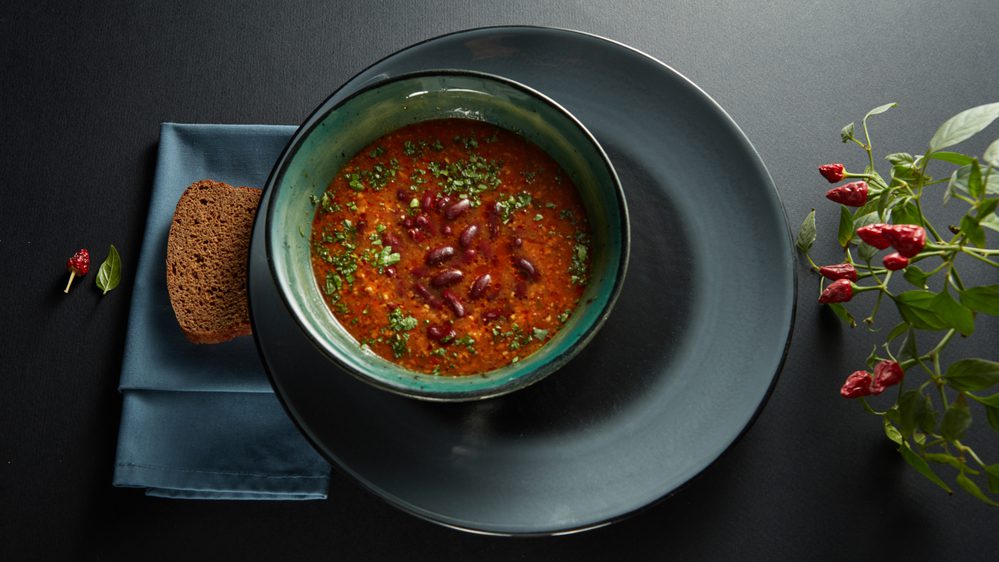 A bowl of bean soup garnished with herbs sits on a dark plate next to a slice of bread and a small chili pepper plant.