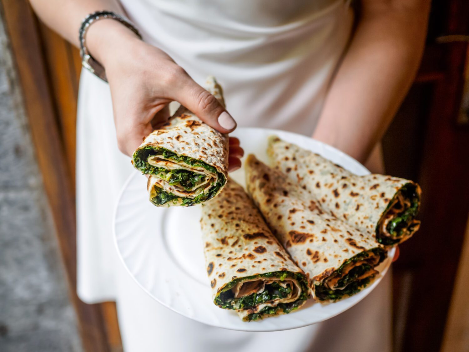 A person in a white dress holds a plate of four rolled flatbreads filled with greens.