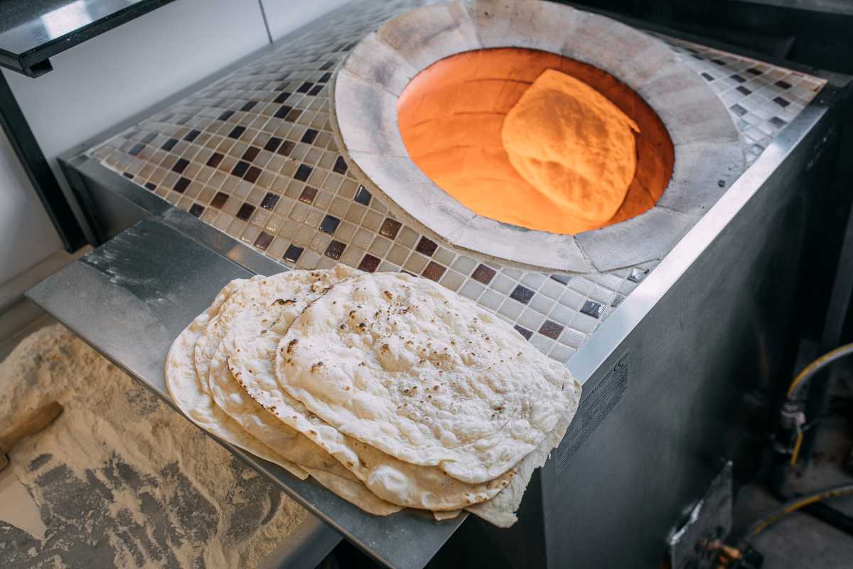 A piece of flatbread bakes inside a tiled oven while a stack of cooked flatbreads rests on a metal surface nearby.