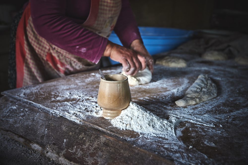 A person in an apron kneads dough on a wooden table next to a small ceramic cup and a pile of flour.