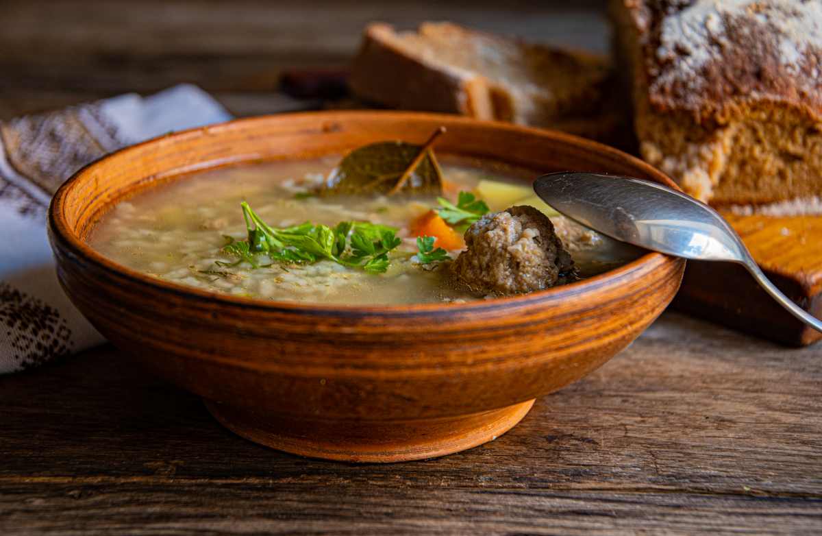 A clay bowl of soup with a meatball, herbs, and a spoon sits on a wooden table next to a slice of dark bread.