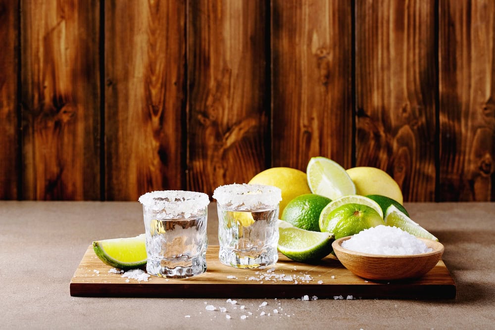 Tequila shots in small glasses with the rims dipped in salt crystals, beside the glasses are a bowl of salt and wedges of lemons and limes
