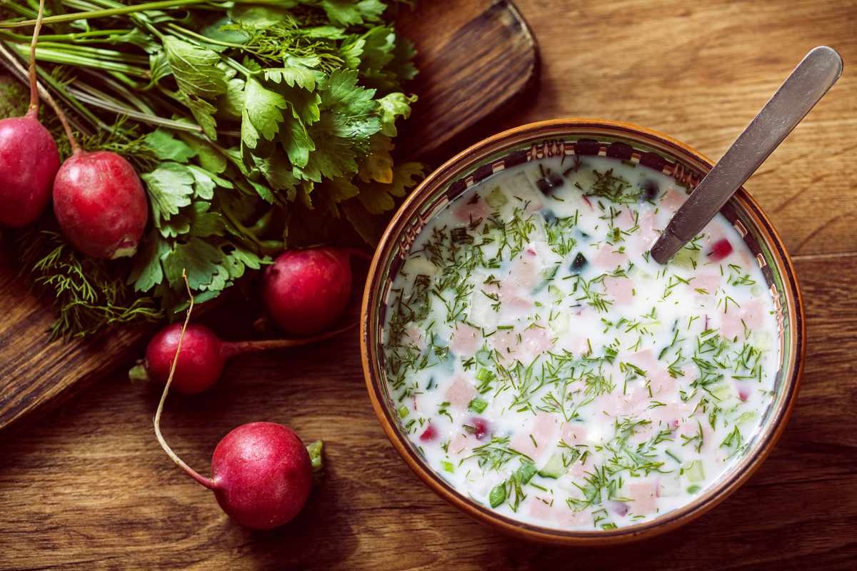 A bowl of cold soup with herbs and diced vegetables sits on a wooden table next to a bunch of fresh radishes.
